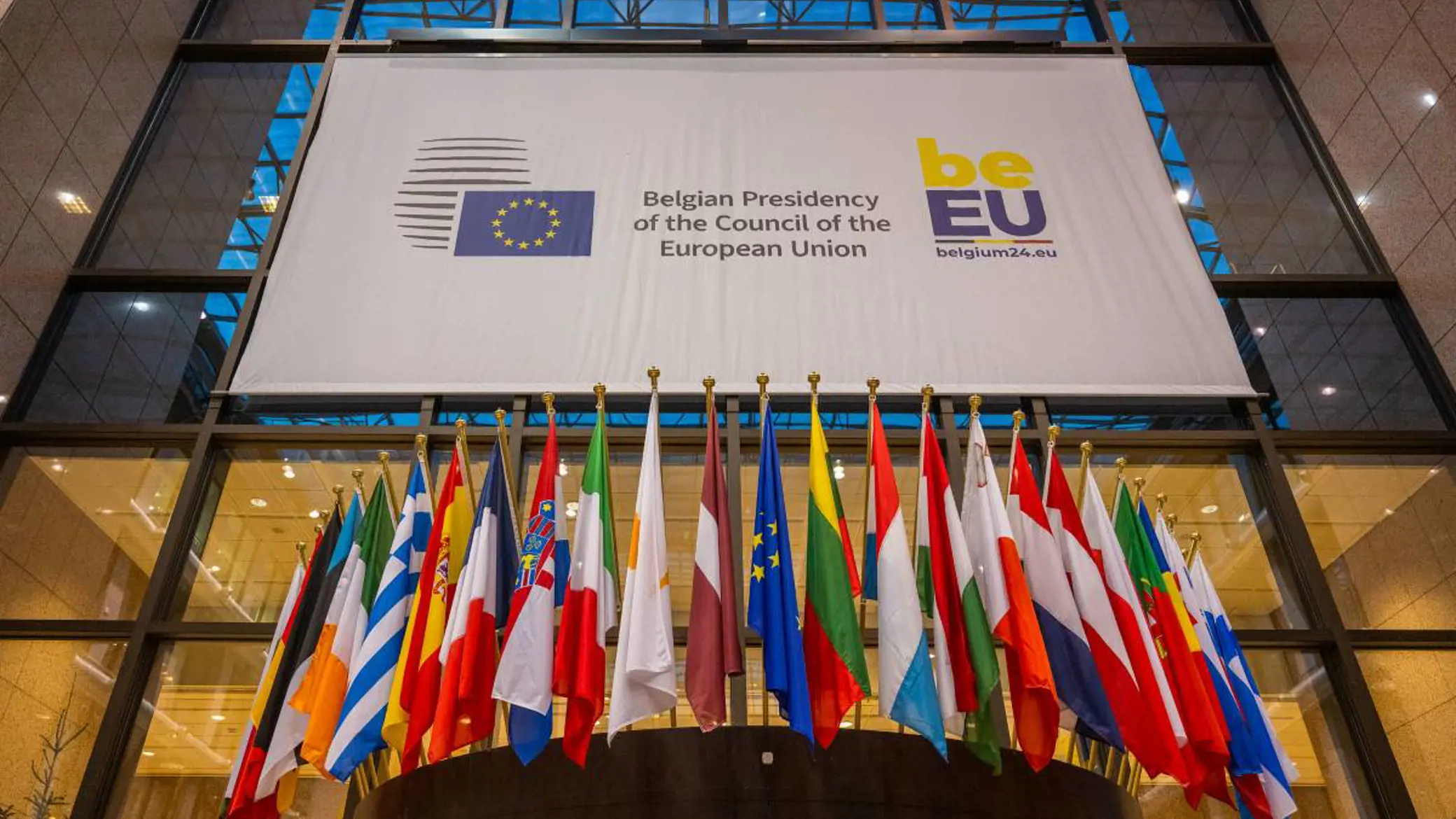 Photo of all 27 flags of the EU inside the main hall of the European Council building in Brussels, under a large banner that states "Belgian Presidency of the Council of the European Union". Photo credit @ Belgian Presidency.